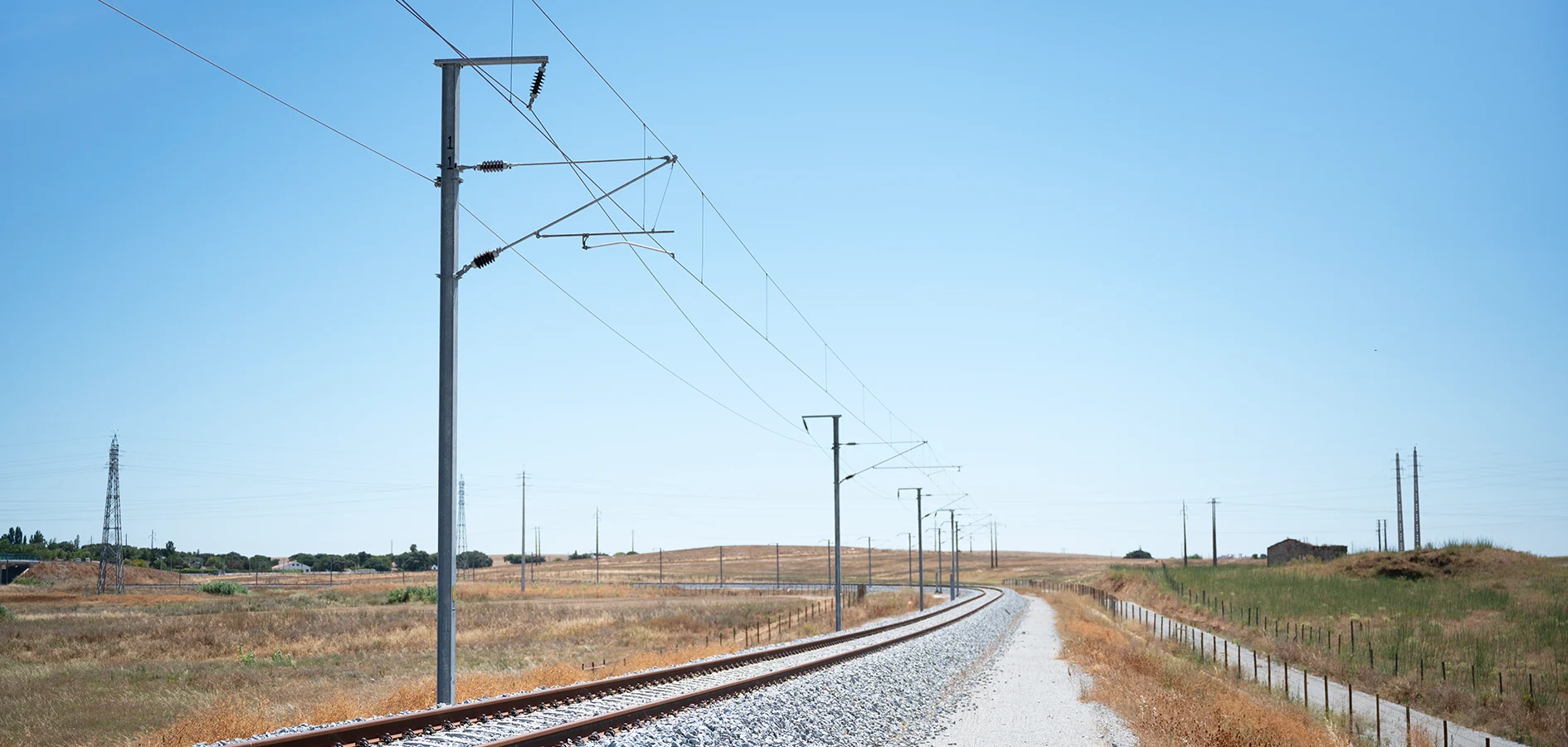 Railway track curving beneath overhead electrification wires and steel support poles.
