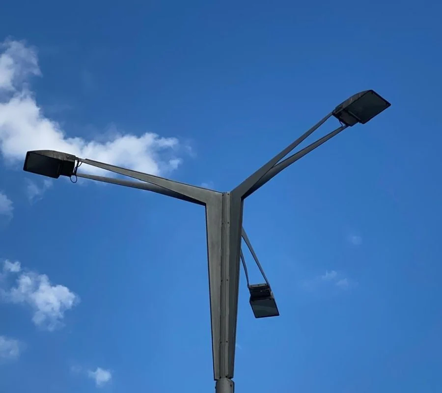 Steel street-lighting pole with three outreach arms against a blue sky.